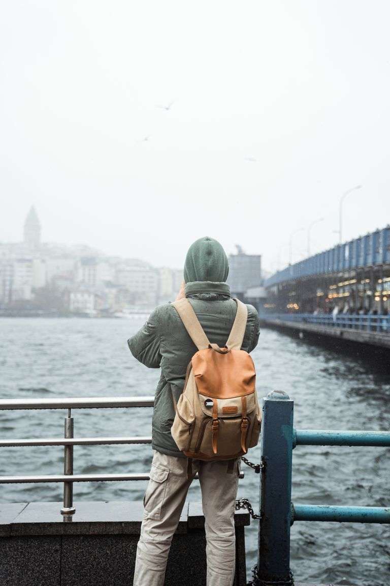 A traveler stands by a riverside railing, gazing at an urban skyline on a cloudy day.