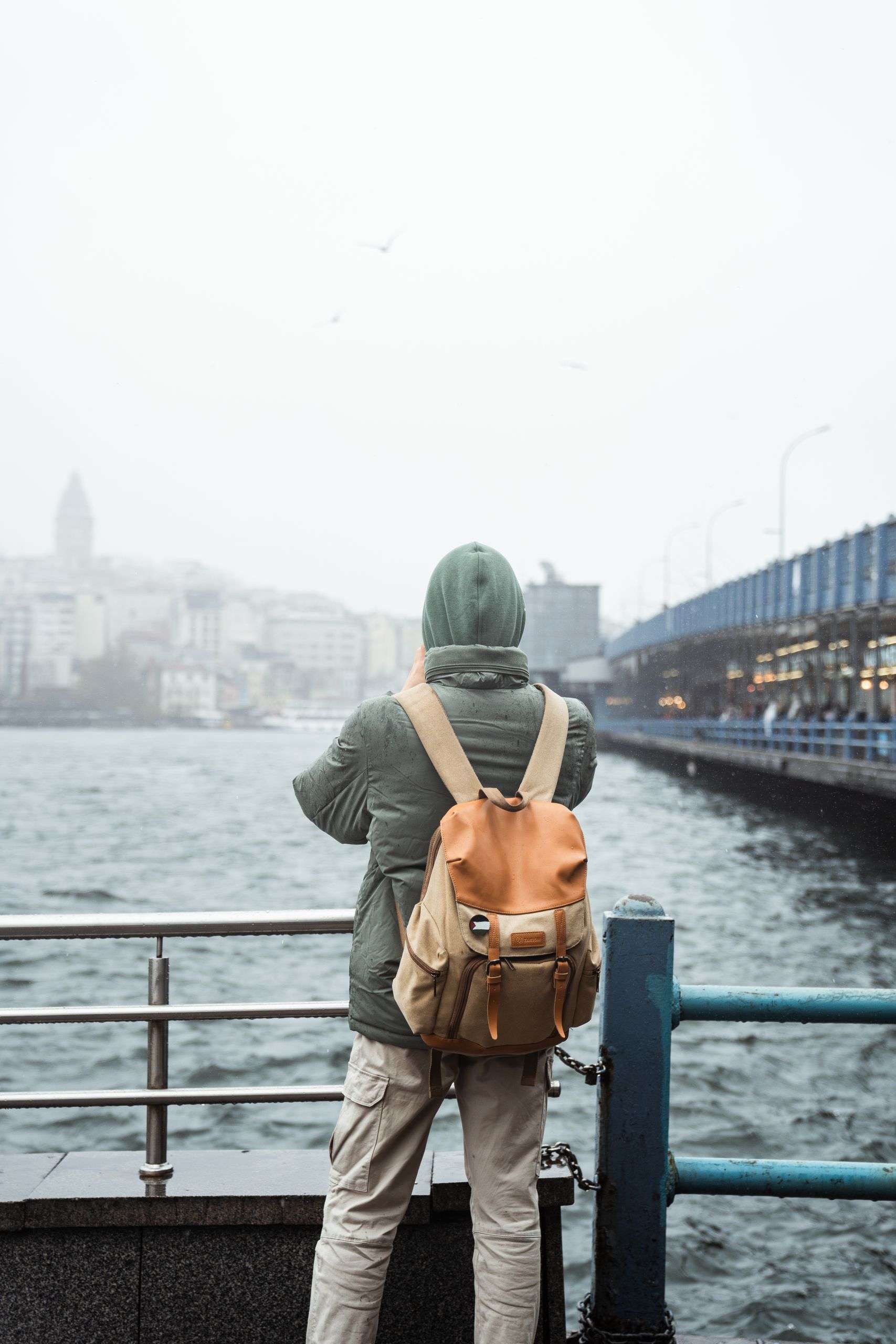 A traveler stands by a riverside railing, gazing at an urban skyline on a cloudy day.