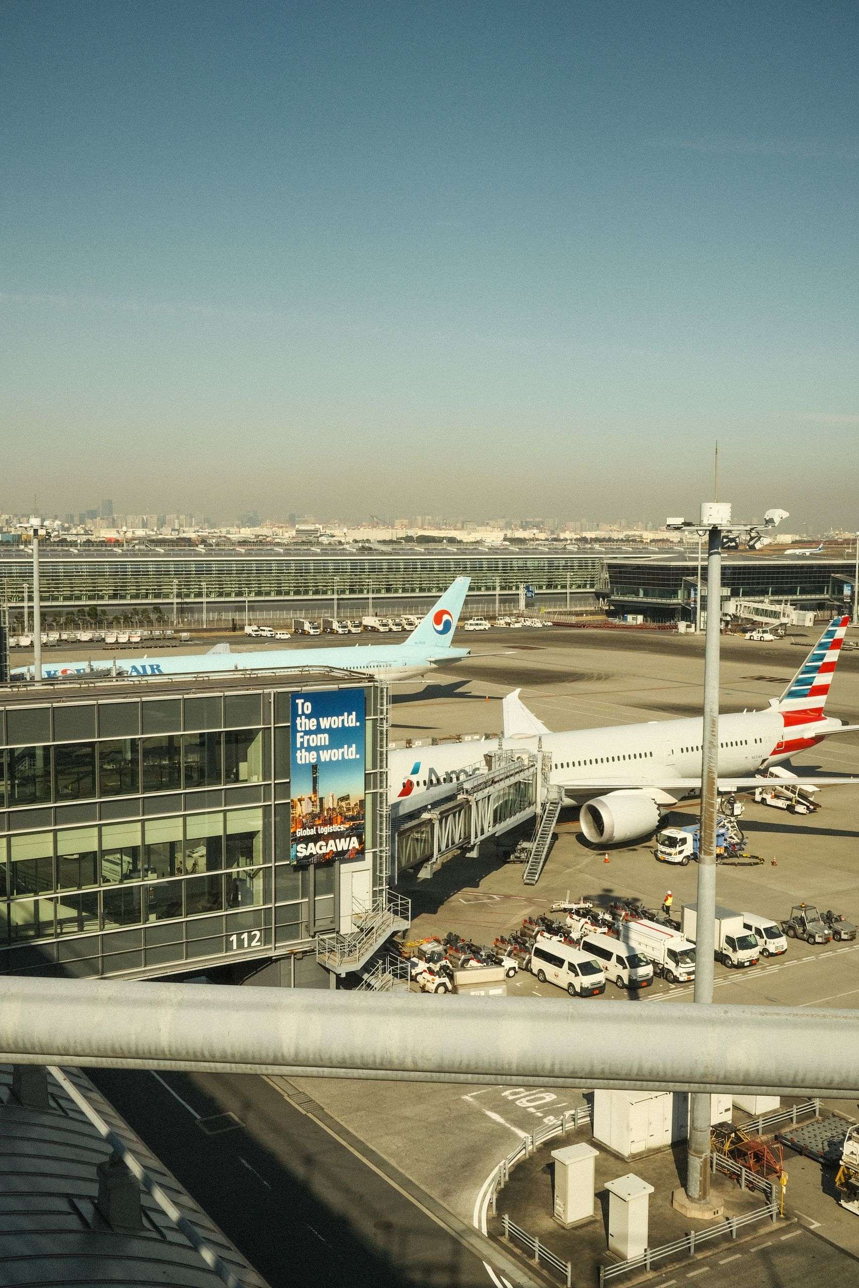 Aerial view of a bustling airport tarmac with parked airplanes and terminal building under clear skies.